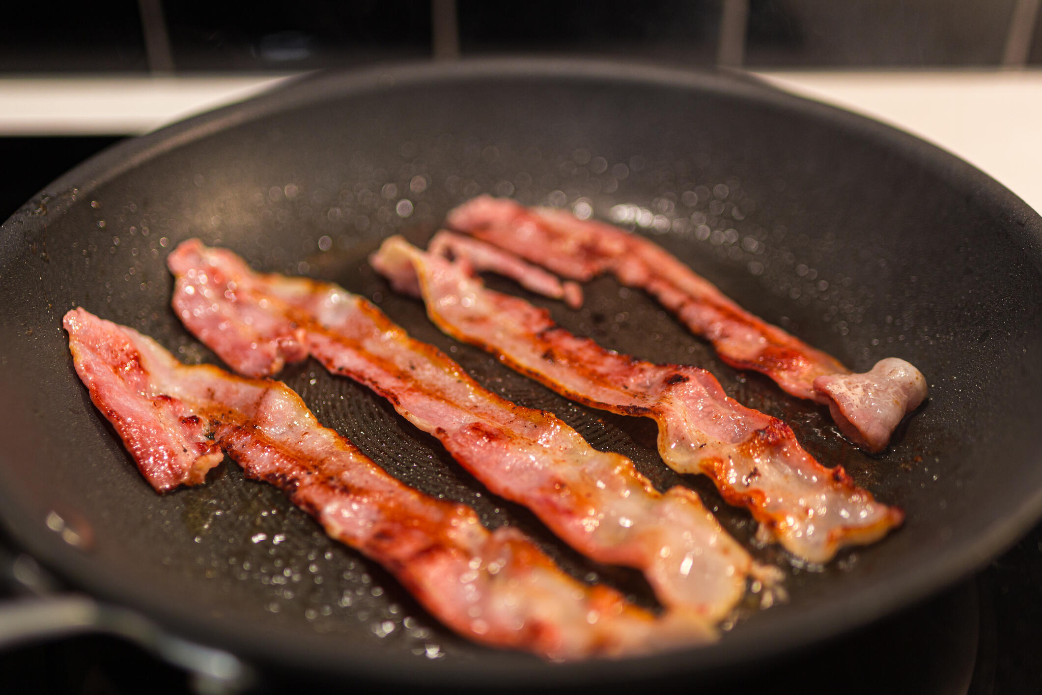 Strips of bacon cooking in a greasy black pan on the stove.