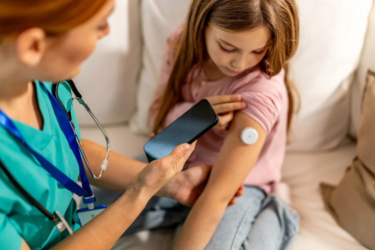 Foto colorida de criança com dispositivo medidor de glicose colad no braço enquanto médica a examina, - Metrópoles.