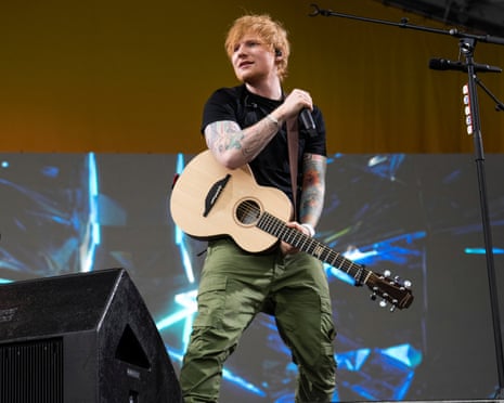 Ed Sheeran sings into a microphone while holding an acoustic guitar on stage