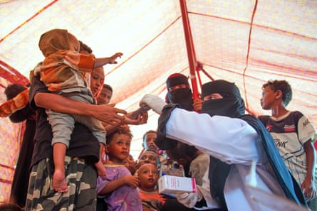 A view inside a large tent of a woman holding a crying baby being given a packet of medicine by an aid worker