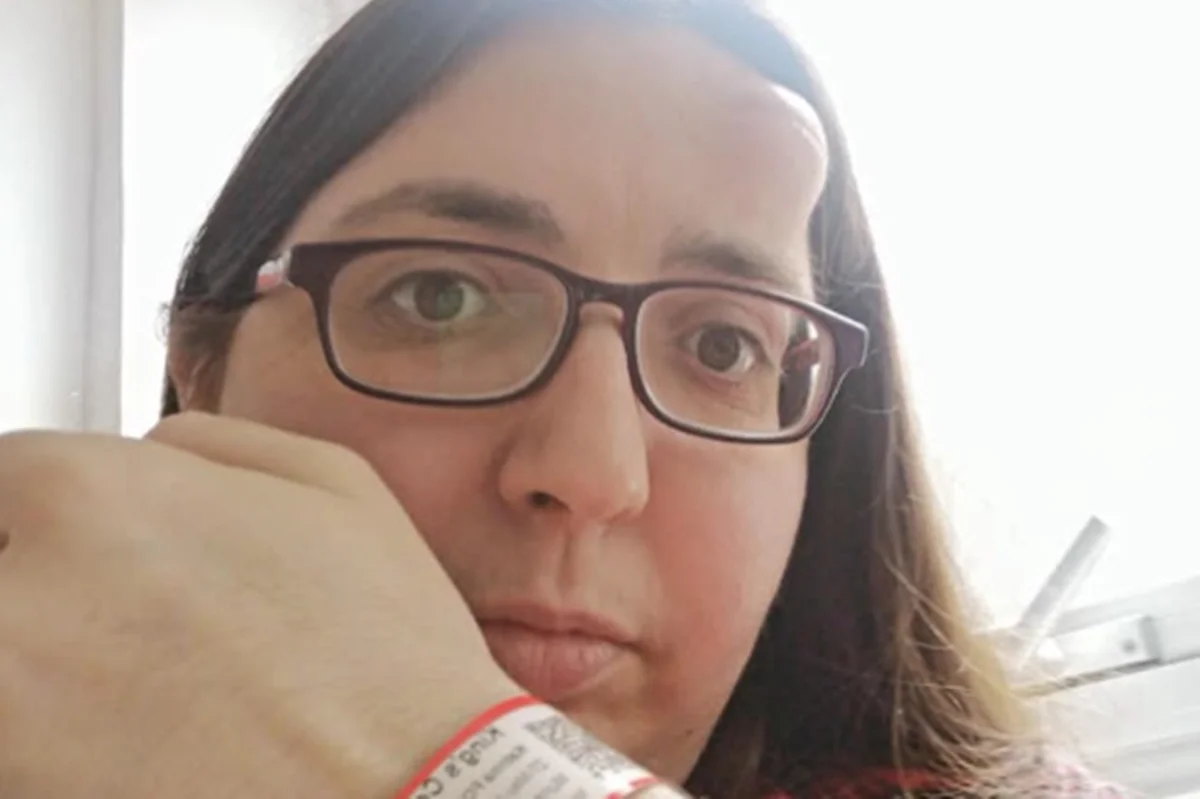 Foto colorida de mulher usando óculos, exibindo uma pulseira de identificação médica, com um caroço enorme na sua testa - Metrópoles.