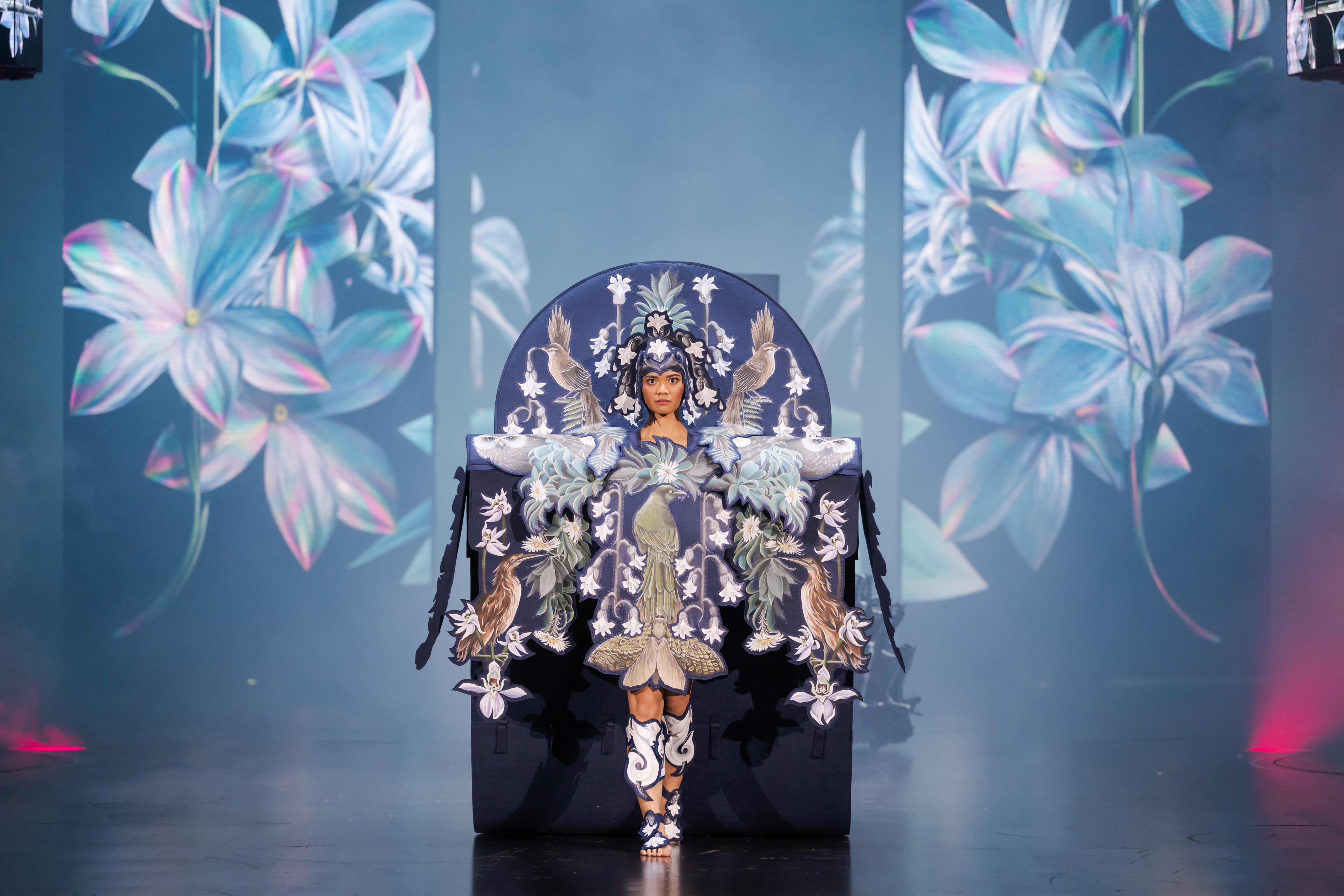 A dancer wears a detailed blue and white costume and headpiece with floral and feather motifs.