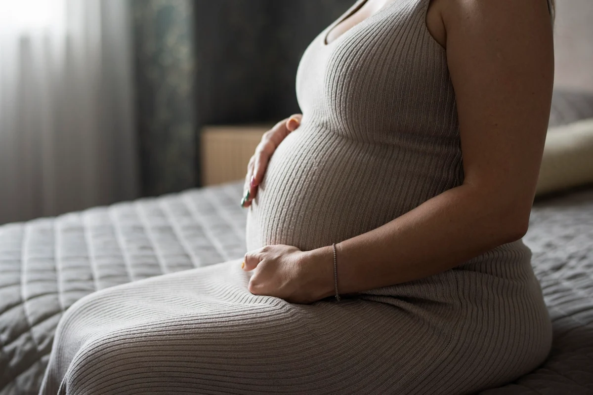Foto colorida de mulher grávida sentada na cama, com close na barriga - Metrópoles.