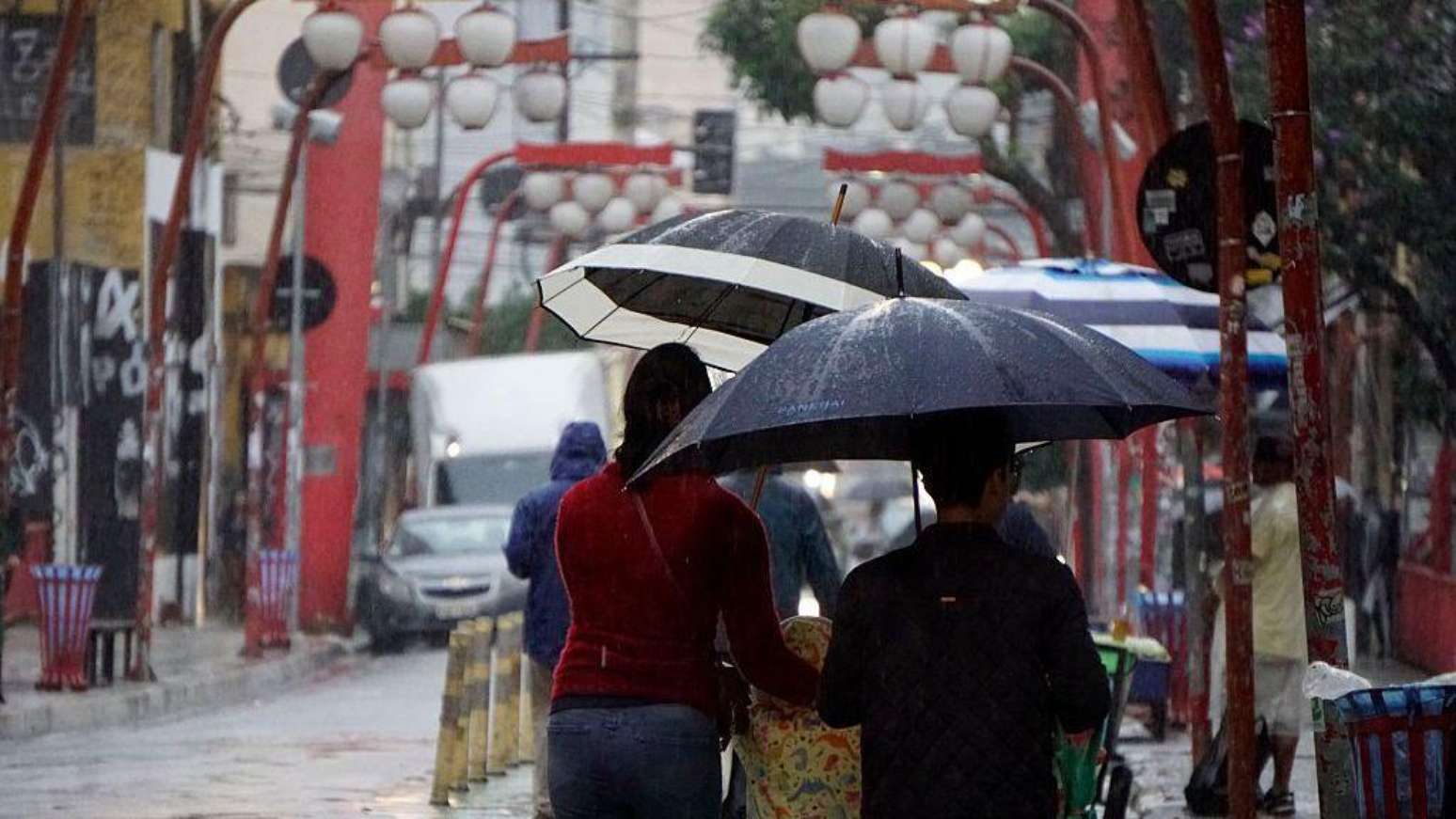 Uma fotografia retangular, tirada ao nível dos olhos, mostrando uma rua movimentada em um dia chuvoso no bairro da Liberdade, em São Paulo. À esquerda, uma mulher veste uma blusa de manga comprida vermelha e calça jeans escura. Ela segura um guarda-chuva aberto de cor preta por fora e branca por dentro. À direita, um homem segura um grande guarda-chuva totalmente preto, inclinado sobre a sua cabeça.