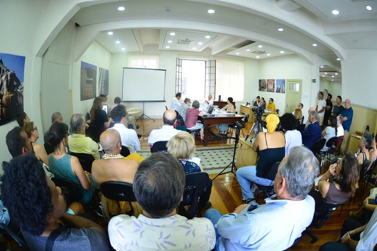 Imagem colorida com lente grande angular mostra uma sala repleta de pessoas. Parte está em uma mesa de reuniões e parte está sentada em cadeiras, na plateia. É uma reunião do Conpresp - Metrópoles