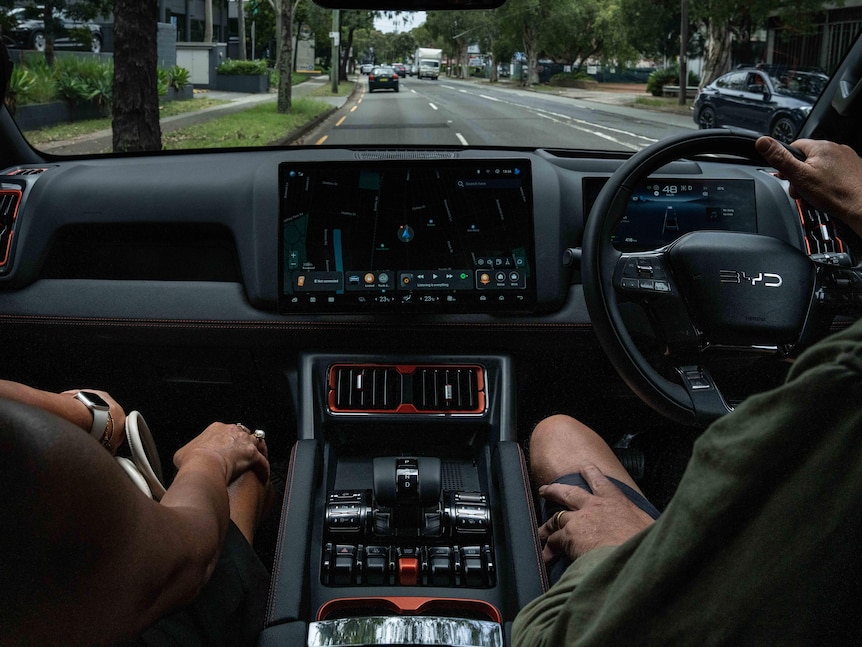 A driver and passenger sit in a modern BYD car driving on the road. The photo is taken from the back seat.